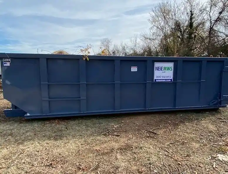 A large blue dumpster placed on bare ground with some vegetation in the background.